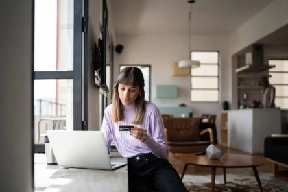 Young woman holding credit card while using laptop at home fg trade.jpg
